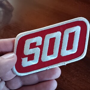 May include: A rectangular patch with the number "500" embroidered in white on a red background. The patch has a white border and is held in a hand.