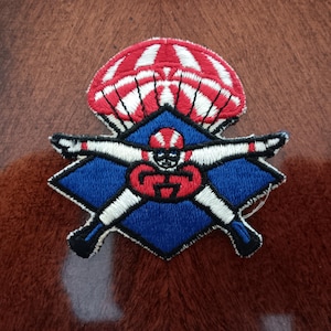 May include: Embroidered patch featuring a skydiver design. The patch has a red and white parachute above a blue diamond shape. Inside the diamond is a skydiver with outstretched arms, wearing a red helmet and uniform. The patch is detailed with white, black, and red stitching.