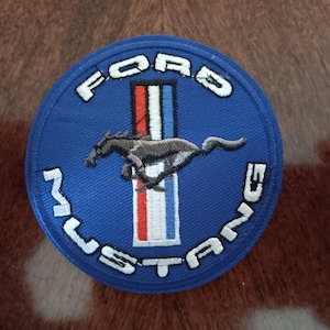 May include: A round, blue embroidered patch featuring the Ford Mustang logo. The patch has white text that reads "FORD MUSTANG" around the edge. A running horse is in the center, above a vertical stripe of red, white, and blue.