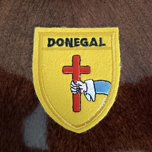 May include: A yellow patch with the word "DONEGAL" in black lettering. The patch features a red cross held by a white-gloved hand with a blue sleeve. The patch is bordered by a yellow stitched edge.