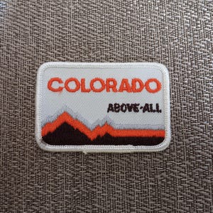 May include: Rectangular patch with the word "COLORADO" in orange block letters above the words "ABOVE ALL". The patch features a mountain range design in orange, gray, and brown. The patch has a white border.