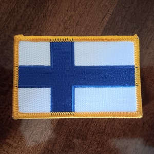 May include: A blue and white embroidered patch with a yellow border, depicting the flag of Finland.