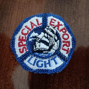 May include: Embroidered patch with a nautical theme. The patch is a hexagon shape with a blue border and features the words "SPECIAL EXPORT LIGHT" in red and white. A detailed image of a ship is in the center.