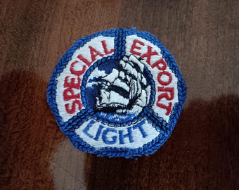 May include: Embroidered patch with a nautical theme. The patch is a hexagon shape with a blue border and features the words "SPECIAL EXPORT LIGHT" in red and white. A detailed image of a ship is in the center.