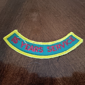 May include: A green, curved patch with yellow trim and red embroidered text that reads "15 YEARS SERVICE". The patch is likely for a service organization or club, and is designed to be sewn onto clothing.
