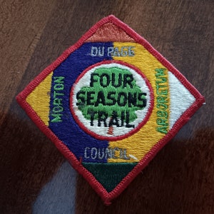 May include: A diamond-shaped patch with a red border. The center features a green tree with the text "Four Seasons Trail" in white. The surrounding text reads "Morton Arboretum, Du Page Council".