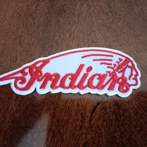 May include: A white embroidered patch featuring the red script text Indian with a stylized red graphic of a person wearing a feathered headdress integrated into the design. The patch has a white border and a textured fabric surface.