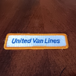 May include: A rectangular patch with the text "United Van Lines" in blue letters. The patch has a white background with a gold border. The patch is likely an emblem or logo.