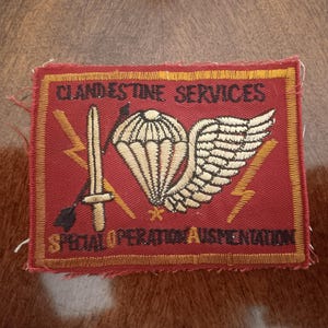 May include: A rectangular red patch with gold embroidered text and imagery. The text reads "CLANDESTINE SERVICES" and "SPECIAL OPERATION AUSMENTATION". The patch features a parachute, sword, lightning bolts, and a wing.