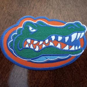 May include: An embroidered patch featuring a green alligator head with sharp white teeth, set against an orange oval background, all bordered by a thick blue stitched outline. The design displays a snarling expression with visible eyes.