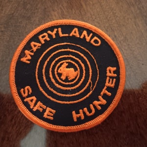 May include: Orange and black embroidered patch with the text "Maryland Safe Hunter" and a stylized rabbit inside a target.