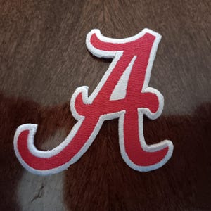 May include: A red embroidered letter "A" patch with a white border. The patch is set against a brown and white textured background. The letter is in a script font.