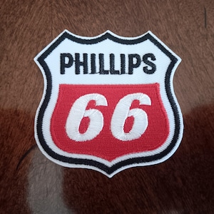 May include: A shield-shaped embroidered patch featuring a white background with a black border. The word PHILLIPS is stitched in black at the top, and the number 66 is stitched in white against a red background in the lower section of the patch.