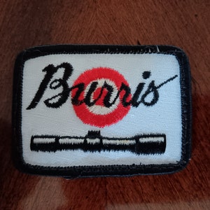May include: A white embroidered patch with black stitching. The patch features the word "Burris" in a cursive font with a red and white target in the center. A black and white illustration of a rifle scope is below the target.