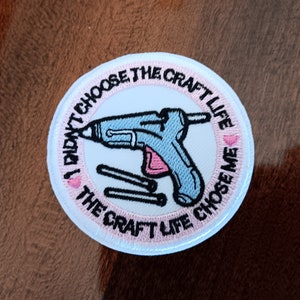 May include: A round, white embroidered patch with the text "I DIDN'T CHOOSE THE CRAFT LIFE CHOSE ME". The patch features a light blue and pink glue gun and three glue sticks. The text and design are outlined in black.