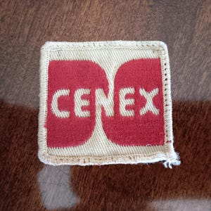 May include: A rectangular patch with the word "CENEX" in white letters on a red background. The patch has a beige border and is likely made of fabric. The patch is on a brown surface.