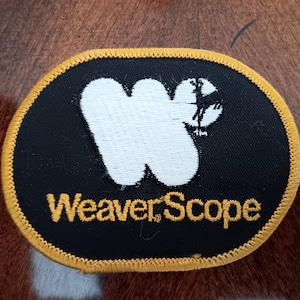 May include: A black and yellow oval-shaped patch with the text "WeaverScope" in yellow embroidery. A white stylized "W" with a deer silhouette is in the center.