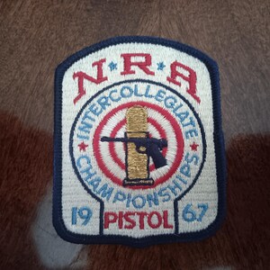 May include: A rectangular embroidered patch with a dark blue border. The patch features the red text NRA, the blue text INTERCOLLEGIATE CHAMPIONSHIPS, and the red text PISTOL with the numbers 19 and 67. A gold bullet and black pistol are centered.