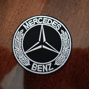 May include: A round, black and white embroidered patch featuring the Mercedes-Benz logo. The logo includes a three-pointed star within a circle, surrounded by the words "MERCEDES" and "BENZ". The patch has a white border and is likely for apparel.