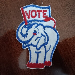 May include: A white embroidered patch of an elephant holding a red, white, and blue flag that says "VOTE".