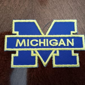 May include: Embroidered patch featuring the letter "M" in blue with a yellow border. The word "MICHIGAN" is embroidered in yellow across the center. This patch is likely for apparel or accessories.
