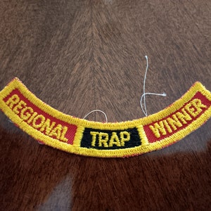 May include: Embroidered patch with the words "REGIONAL TRAP WINNER" in yellow lettering on a red and black background. The patch has a curved shape and a yellow border. A few loose threads are visible.