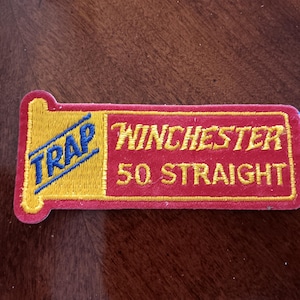 May include: Embroidered patch with the word "TRAP" in blue and yellow, and "Winchester 50 Straight" in gold lettering on a red background. The patch has a rectangular shape with rounded corners.