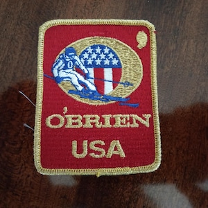 May include: A red and gold embroidered patch with a skier in a blue and white uniform, a US flag, and the text "O'Brien USA".