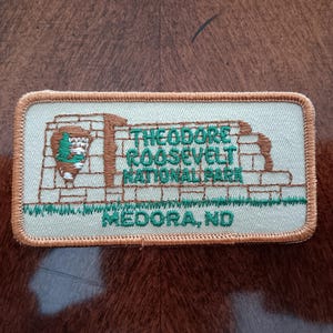 May include: Embroidered patch featuring the text "THEODORE ROOSEVELT NATIONAL PARK MEDORA, ND" in green. The patch has a light green background with a brown and green design of a park entrance. The patch is rectangular with a tan border.