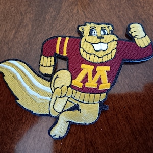May include: Embroidered patch featuring a cartoon gopher mascot in a maroon sweater with gold accents and the letter "M". The gopher is in a running pose with a large, fluffy tail. The patch is outlined in black.