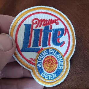 May include: A circular embroidered patch featuring the Miller Lite logo in blue and red text on a white background with a gold border. Below the main logo, a smaller circular emblem contains the text A TRUE PILSNER BEER in blue.
