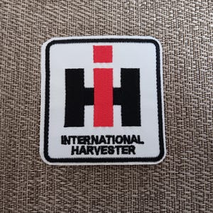 May include: A square embroidered patch featuring the International Harvester logo. The design includes a large black letter H with a red letter i centered vertically, and the text INTERNATIONAL HARVESTER in black below, all on a white background.