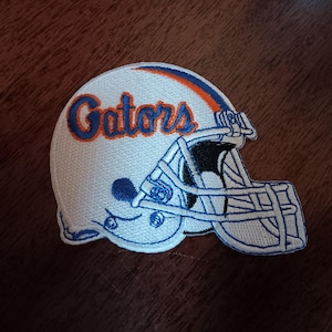 May include: Embroidered patch featuring a white football helmet with blue and orange accents. The word "Gators" is stitched in blue and orange on the helmet. The patch is likely for apparel or accessories.