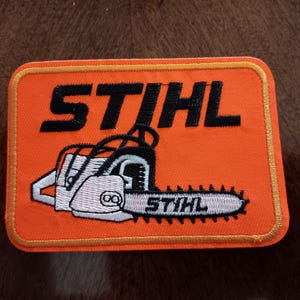 May include: A rectangular embroidered patch featuring a bright orange background with a gold-colored border. The text STIHL is stitched in black at the top, and a white and black chainsaw graphic with the word STIHL on the bar is centered below.