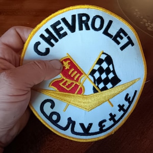 May include: Embroidered patch featuring the Chevrolet Corvette logo. The patch is white with black text that reads "CHEVROLET" and "Corvette". It includes a red and gold emblem and a checkered flag.
