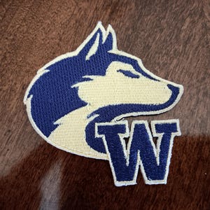 May include: An embroidered fabric patch featuring a stylized wolf head in dark blue and cream colors, positioned above a large, bold, dark blue capital letter W with a cream-colored border, creating a cohesive graphic design element.