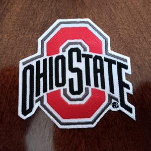 May include: An embroidered patch featuring the Ohio State logo in red, white, black, and grey thread. The design includes the word OHIO STATE in bold black lettering layered over a large red block letter O with white and grey borders.