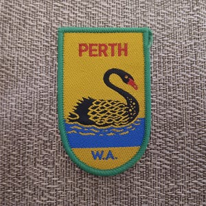 May include: A patch featuring a black swan with a red beak on a yellow background, with the word "PERTH" in red at the top and "W.A." in blue at the bottom. The patch has a green border.