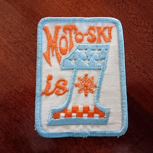 May include: Embroidered patch with the words "Moto-Ski is 1" in orange script. The design includes a blue and orange number one with a snowflake and mountain graphic. The patch has a white background and a blue border.