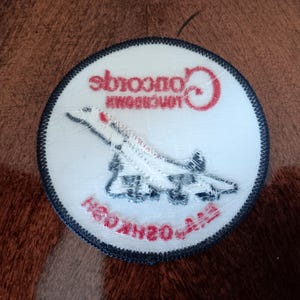 May include: A circular embroidered patch with a white background and a dark blue border. It features a white Concorde airplane graphic, the red text Concorde, and the red text EAA OSHKOSH printed in a curved orientation at the bottom.