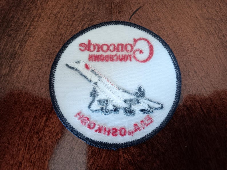 May include: A circular embroidered patch with a white background and a dark blue border. It features a white Concorde airplane graphic, the red text Concorde, and the red text EAA OSHKOSH printed in a curved orientation at the bottom.
