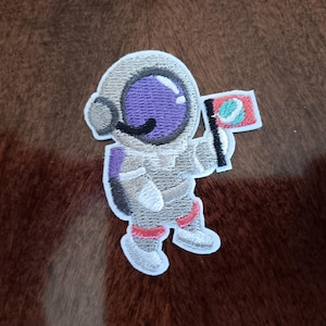 May include: Embroidered patch featuring a cartoon astronaut in a spacesuit, holding a flag. The astronaut's helmet has a purple visor, and the flag is red with a blue and green design. The patch is gray, white, purple, red, and black.