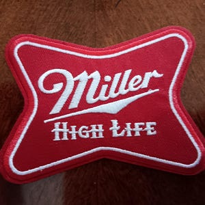 May include: A red, cross-shaped embroidered patch with a white border. The patch features the white embroidered text Miller and High Life, with the Miller logo positioned above the words High Life in a stylized, bold font on a red fabric background.