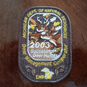 May include: Embroidered patch featuring a deer scene with the text "2003 Successful Deer Hunter". The patch is brown with a white border and includes the text "Michigan Dept. of Natural Resources".