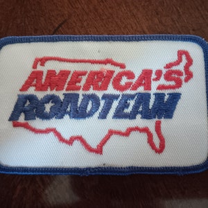 May include: A white embroidered patch with a blue outline. The patch features a red outline of the United States and the text "America's Roadteam" in red and blue lettering.