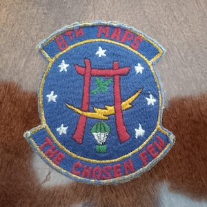 May include: Embroidered patch with a blue background, featuring red archways, white stars, and yellow lightning bolts. The text "8TH MARS" and "THE CHOSEN FEW" are in red. A green and white parachute is also visible.