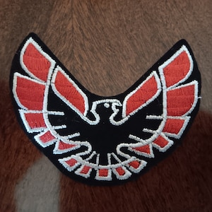 May include: Black embroidered patch featuring a stylized eagle design with red and white accents. The patch has a curved shape and is likely for application on clothing or accessories. The eagle is the central focus.