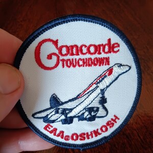 May include: A circular embroidered patch with a white background and a dark blue border. It features a red embroidered Concorde airplane graphic, the red text "Concorde TOUCHDOWN" at the top, and the red text "EAA OSHKOSH" at the bottom.