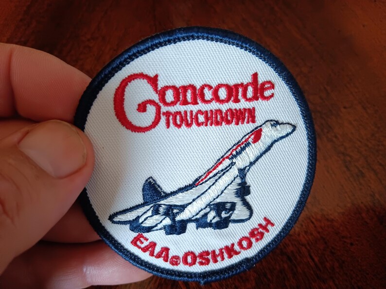 May include: A circular embroidered patch with a white background and a dark blue border. It features a red embroidered Concorde airplane graphic, the red text "Concorde TOUCHDOWN" at the top, and the red text "EAA OSHKOSH" at the bottom.