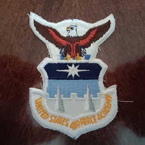 May include: Embroidered patch featuring the United States Air Force Academy emblem. The patch has a white border with the words "UNITED STATES AIR FORCE ACADEMY" in yellow. The emblem includes an eagle, a star, and a stylized building design.